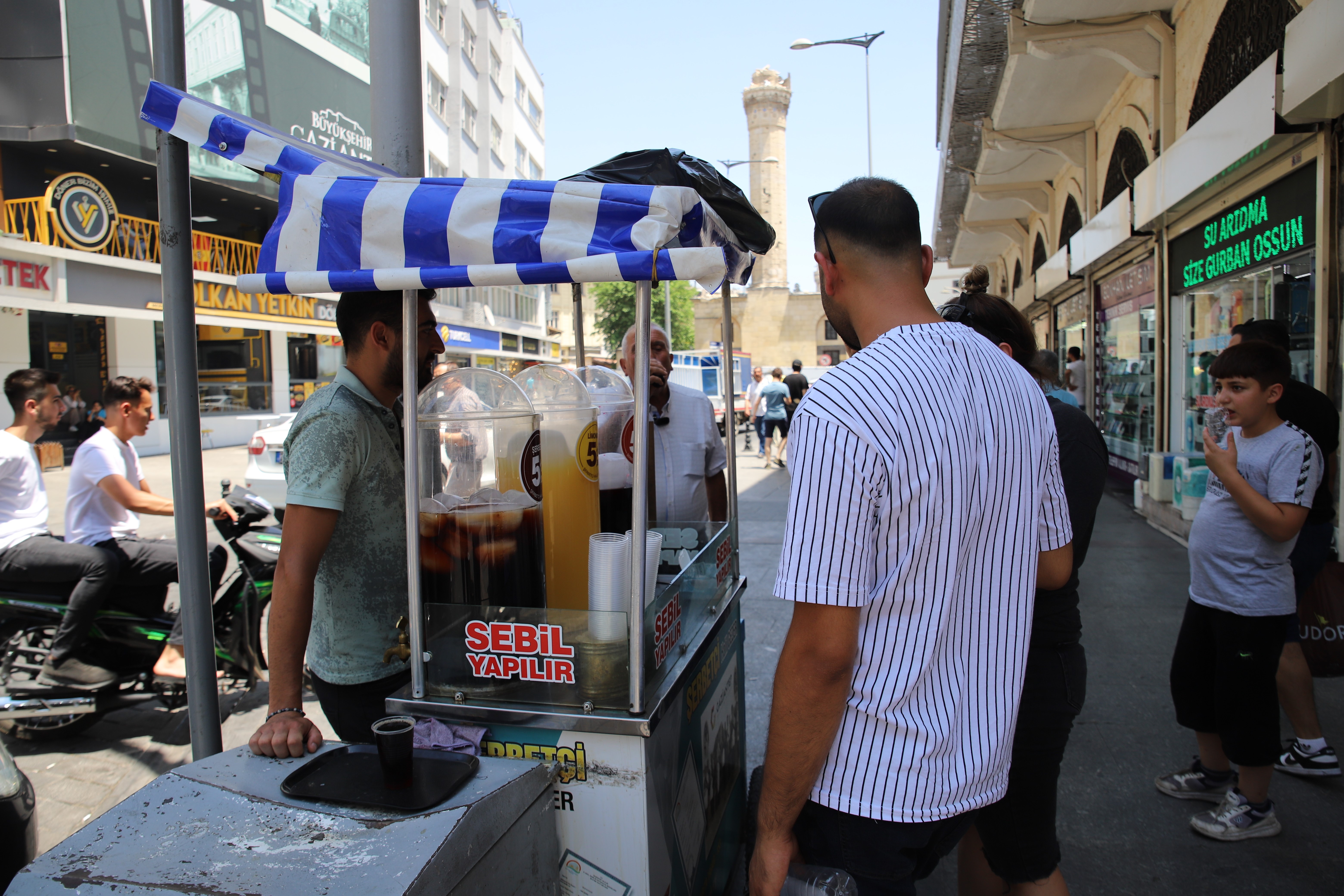 “GAZİANTEPLİ HİÇBİR ŞEKİLDE MEYAN ŞERBETİNDEN BAŞKA HİÇBİR İÇECEĞE TAMAH ETMEZ”
Gaziantep'in 7. nesil meyan şerbeti ustası Bahri Gezer, “Babam Gaziantep’in 6. nesil ben ise 7. nesil şerbetçisiyim. Yazın sıcak günlerinde meyan şerbeti, limonata, vişne suyu meşrubat çeşitlerimizle halkımıza hizmet ediyoruz. Havalar sıcak olduğundan dolayı limonata, meşrubat ve şerbetlere yoğun bir talep var. Kaliteden de ödün vermediğimiz için sık sık müşterilerimiz geliyor. Havalar sıcak olduğundan dolayı meyan şerbeti doğal, yüzde 100 hiçbir katkı maddesi olmadığı için genelde bilen insanlar şerbet içiyor. Hem mideyi rahatlatması ve hazmı kolaylaştırması açısından şerbeti tercih ediyorlar. Daha önce bilmeyen hiç içmemiş olanlar şehir dışından gelenler ise limonata ve vişne suyunu tercih ediyorlar. Gazianteplilerin içeceği meyan şerbetidir. Ramazan sofralarından da anlaşılacağı üzere vazgeçilmez içeceğidir. Gaziantepli doğup büyüyen biri hiçbir şekilde meyan şerbetinden başka hiçbir içeceğe tamah etmez. Tabi yabancılar çok aşina olmadığı için ilk içişte de acı tadı olduğundan dolayı meşrubat çeşitlerine yöneliyorlar” dedi.